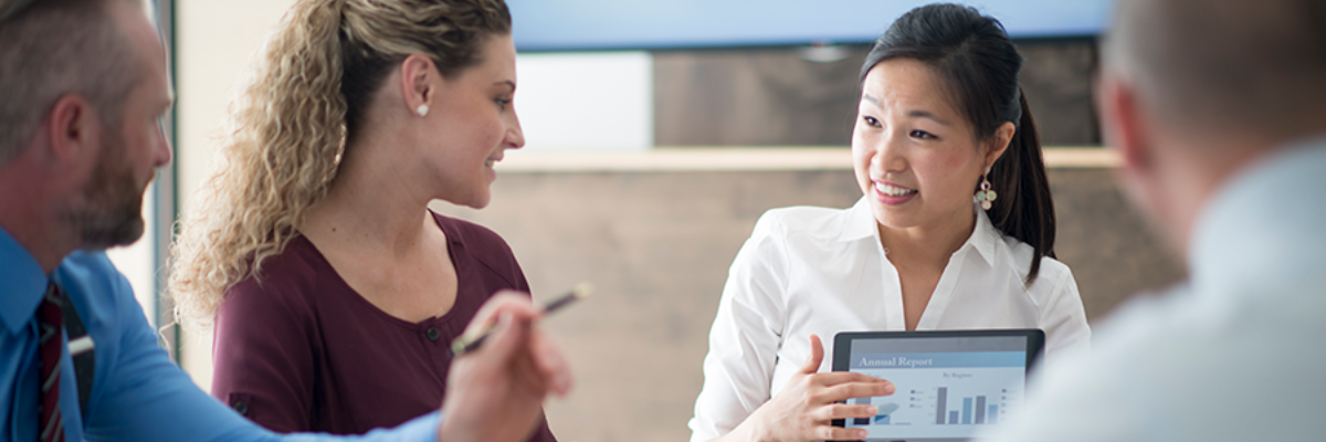 two women, and two men in a meeting looking at a tablet with charts on it. 